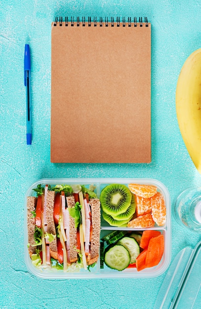 Free photo school lunch box with sandwich, vegetables, water, and fruits on table.