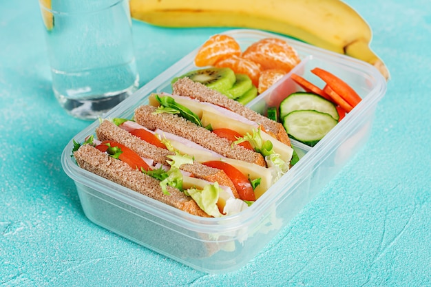 Free photo school lunch box with sandwich, vegetables, water, and fruits on table.