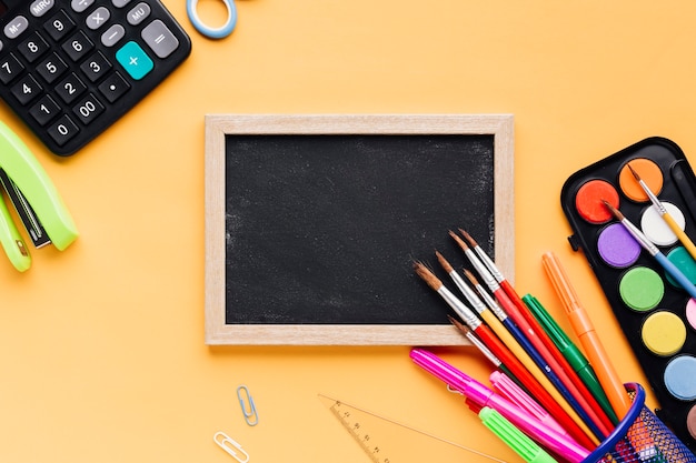 School supplies scattered round blank framed chalkboard on yellow desk