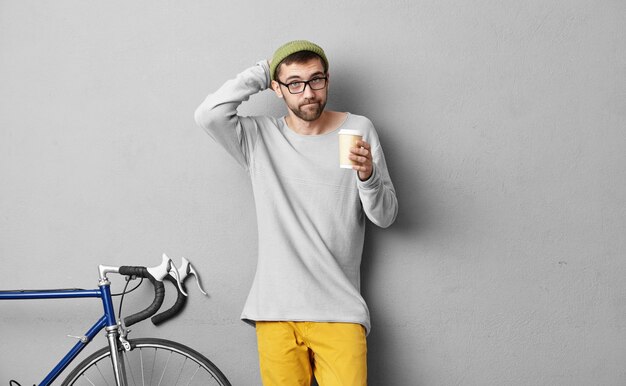 Schoolboy stratching his head while keeping takeaway coffee, trying to remember what he should take in his bag, standing near grey wall and bicycle. Male going on picnic by bike. People and rest