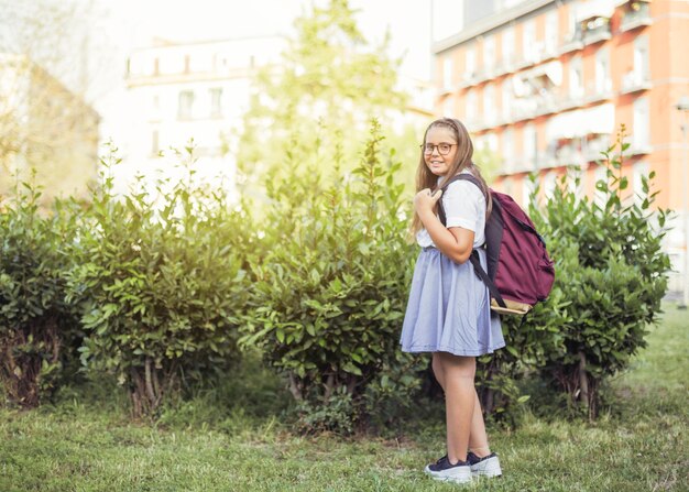 Schoolgirl with backpack standing in front of bushes smiling