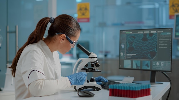 Free photo science engineer using microscope to analyze dna sample in laboratory. woman biologist working with microscopic glass lens on lab tool and computer, wearing protective goggles and gloves.