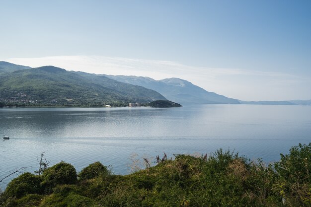 Sea with boats on it surrounded by hills covered in greenery under sunlight