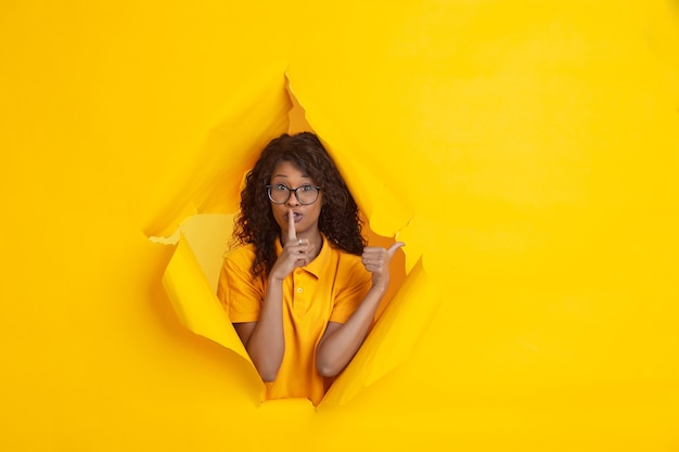 Secretly pointing. Cheerful african-american young woman in torn yellow paper, emotional, expressive.