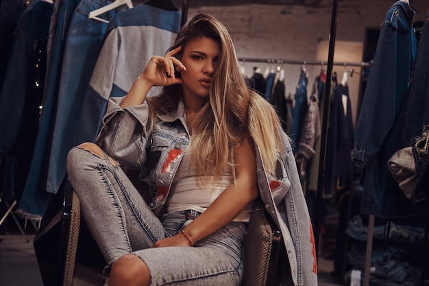 Free Photo seductive girl wearing a distressed jeans and denim jacket looking at a camera while sitting on a chair in the fitting room of a clothing store.