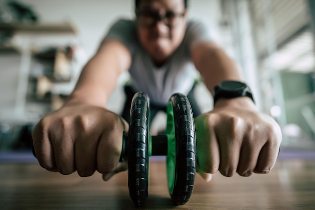 Free photo selective focus funny overweight man wearing eyeglasses is workout with abs handroller for healthy copy space