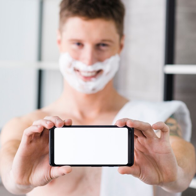 Selective focus of a man with shaving foam on his face showing blank white smart phone toward camera