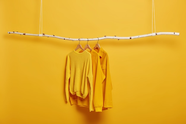 Free photo selective focus. three items of clothes on hangers. long sleeved yellow jumpers on wooden rack near bright vivid wall.