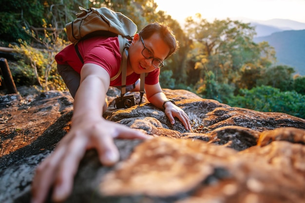 Selective focus Young tourist backpack man wearing eyeglasses trying to climb up the rock for reach the destination trekking trip concept sunlight behind him copy space