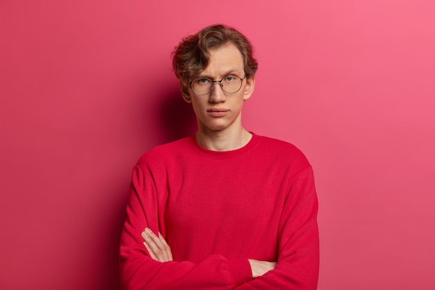 Free photo self assured serious man keeps arms folded, looks directly  with confident expression, has wavy hair, thinks over future plans, wears spectacles and red jumper, isolated on pink wall