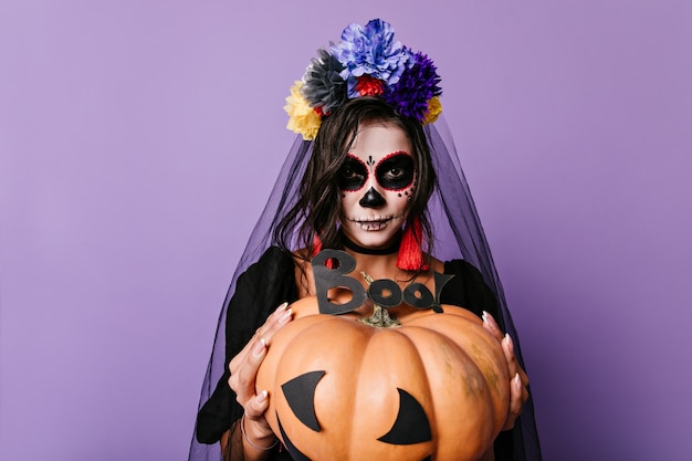 Self-confident girl prepared unusual image and scenery for day of all dead. Photo of woman with bright earrings on isolated wall.