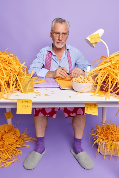 Self confident male director works from home during coronavirus pandemic makes notes sits at messy desktop has breakfast poses at domestic office against purple wall