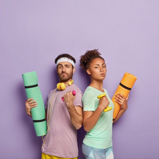 Self confident serious diverse woman and man stand back to each other, raise arms with dumbbells, hold karemats, show their strength, being in good physical shape, isolated on purple  wall