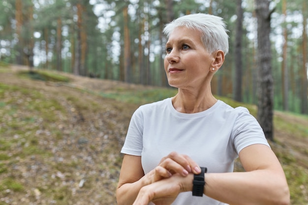 Self determined middle aged sportswoman in white t-shirt adjusting smart watch, checking fitness statistics, monitoring her running performance during cardio workout in park