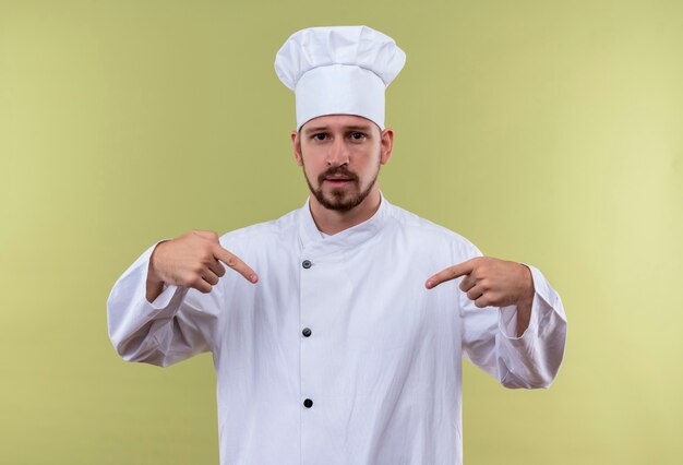 Self-satisfied professional male chef cook in white uniform and cook hat pointing to himself with both hands standing over gree background