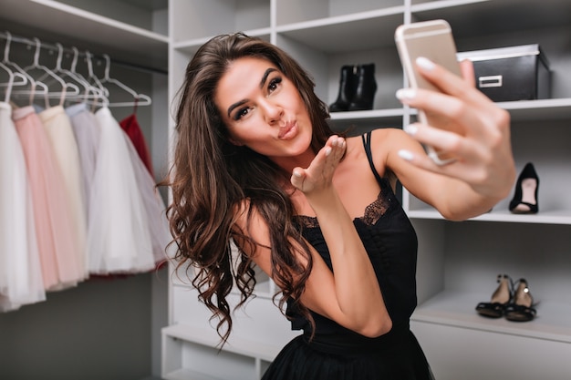 Free photo selfie-portrait of a beautiful brunette girl making a selfie using a smartphone in her dressing room. she sends a kiss. her stylish clothes, expressing true positive face emotions.