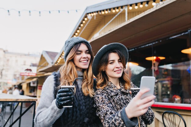 Selfie portrait of joyful fashionable women having fun on sunny street in city. Stylish look, having fun, travelling with friends, smiling, expressing true positive emotions.