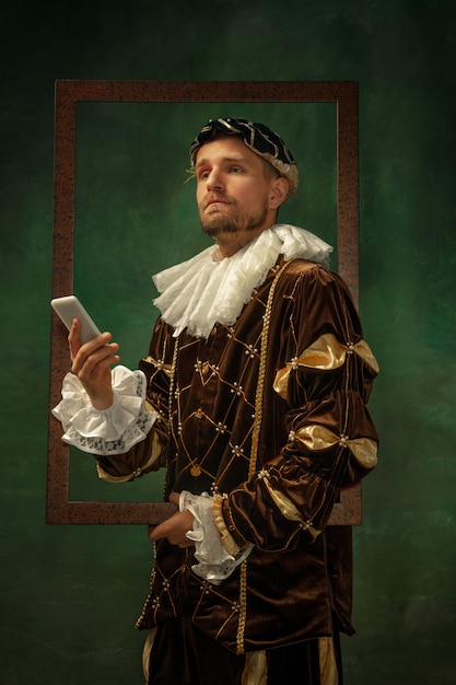 Free photo selfie time. portrait of medieval young man in vintage clothing with wooden frame on dark background. male model as a duke, prince, royal person. concept of comparison of eras, modern, fashion.