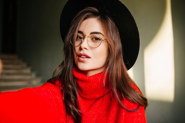 Selfie of young European girl with wavy dark hair in red sweater. Model in hat and glasses posing