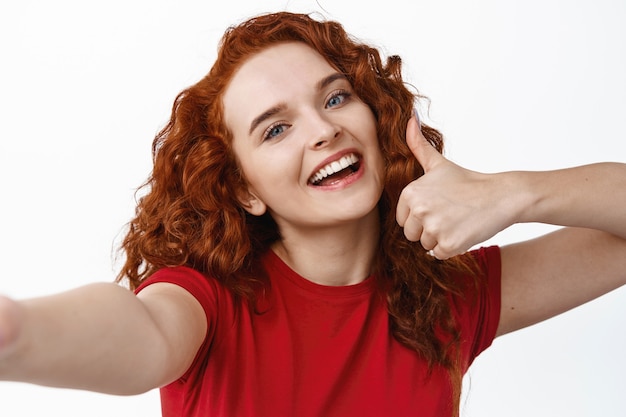 Selfie of young happy redhead girl with curly hairstyle showing thumb up, praise and recommend something good, smiling at smartphone, white wall