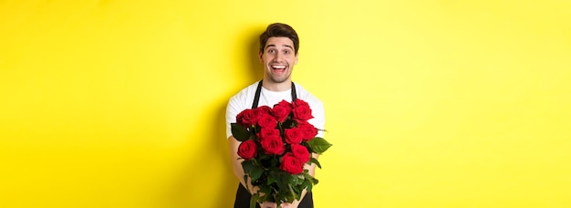 Free photo seller in flower shop wearing black apron giving bouquet of roses and smiling standing over yellow b