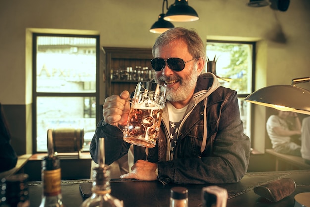 Senior bearded man drinking alcohol in pub and watching a sport program on TV. Enjoying my favorite teem and beer. Man with mug of beer sitting at table. Football or sport fan. Human emotions concept