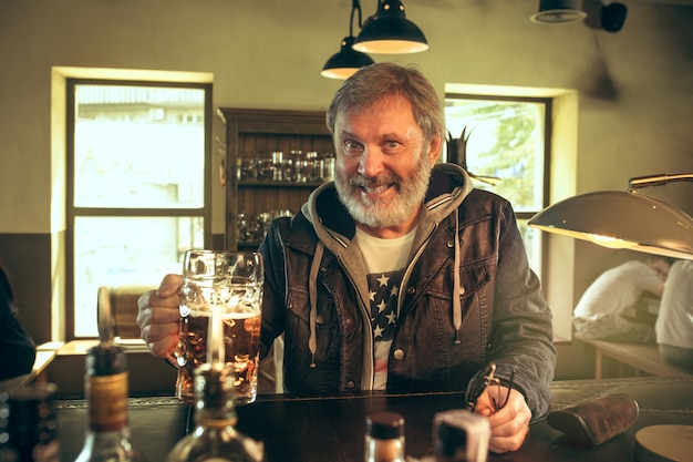 Senior bearded man drinking alcohol in pub and watching a sport program on TV. Enjoying my favorite teem and beer. Man with mug of beer sitting at table. Football or sport fan. Human emotions concept