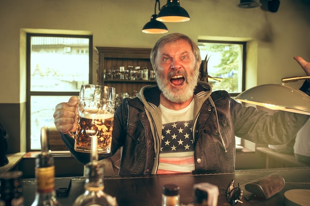 Free Photo senior bearded man drinking alcohol in pub and watching a sport program on tv. enjoying my favorite teem and beer. man with mug of beer sitting at table. football or sport fan. human emotions concept