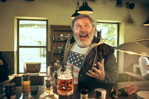 Free Photo senior bearded man drinking alcohol in pub and watching a sport program on tv.