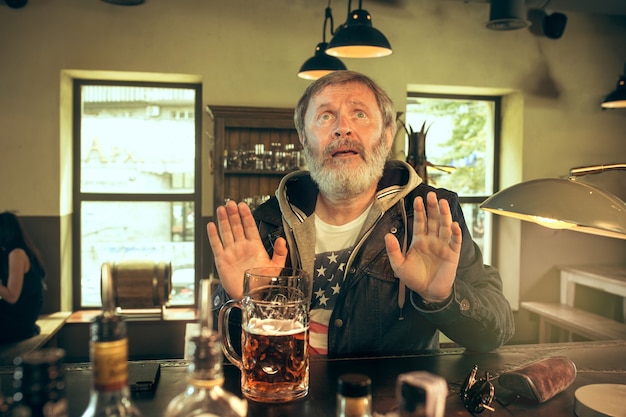 Free photo senior begging bearded man drinking alcohol in pub and watching a sport program on tv. enjoying my favorite teem and beer. man with mug of beer sitting at table. football or sport fan. human emotions