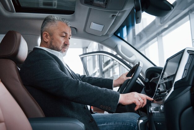 Senior businessman in official clothes sits in a luxury car and pushing the buttons on the music player