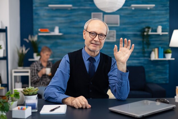 Senior businessman waving at camera wearing glasses during video call