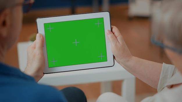 Senior couple holding digital device with green screen at home. Elder husband and wife using mock up template with blank chroma key and isolated copy space on display background. Close up