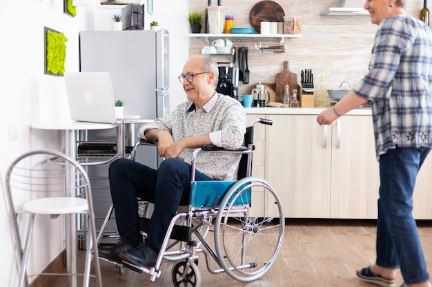 Free Photo senior couple laughing using laptop, husband calling his wife near him during a video call with grandchildrens sitting in kitchen. paralyzedhandicapped old elderly man using communication techonolgy