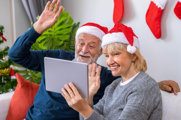 Senior couple using tablet to video phone call to greeting their family for Christmas festival wave hand sitting on sofa with decoration and tree