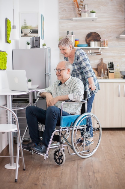 Senior couple on a video call in the kitchen. Disabled senior man in wheelchair and his wife having a video conference on laptop in kitchen. Paralyzed old man and his wife having a online conference.