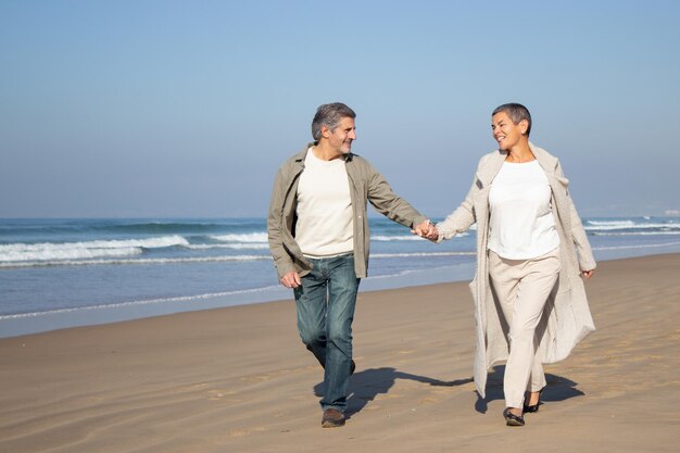 Senior couple walking along seashore on sunny day. Smiling man with gray hair and happy short-haired woman in coat spending time together outdoors. Relationship, partnership concept