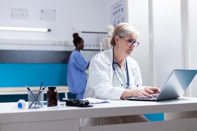 Senior doctor using laptop in cabinet to search for healthcare treatment. General practitioner looking at computer screen to work on prescription medicine against disease diagnosis.