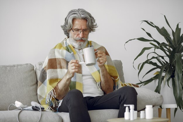 Senior man alone sitting on sofa. Sick man covered with plaid. Grangfather with cup of tea.