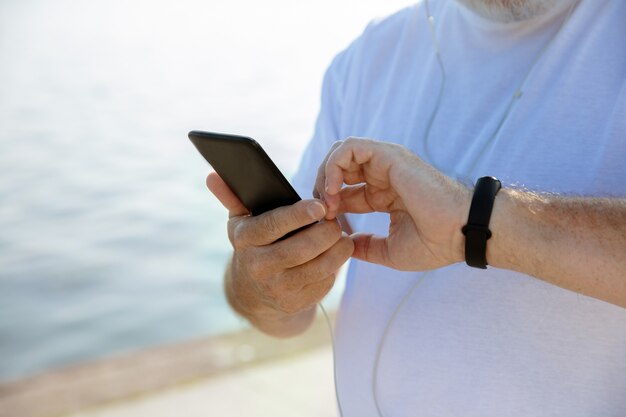 Senior man as runner with fitness tracker at the riverside. Caucasian male model using gadgets while jogging and cardio training in summer's morning. Healthy lifestyle, sport, activity concept.