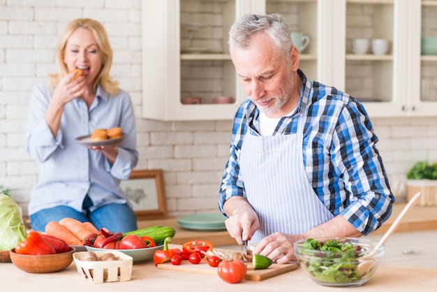 Senior man cutting the vegetables on chopping board with her wife eating the muffins at background in the kitchen