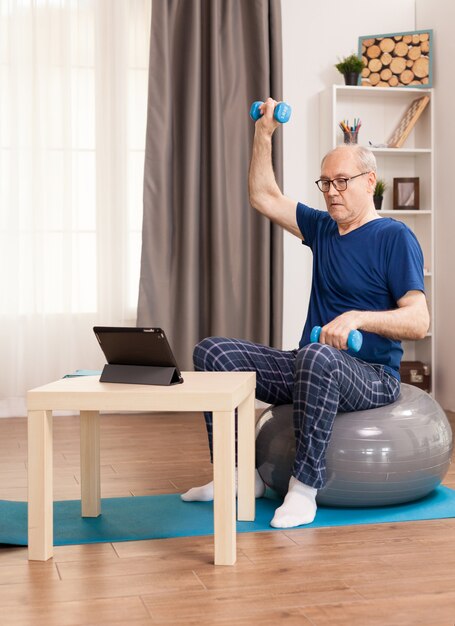 Senior man doing muscle recovery exercises sitting on a swiss ball in front of the tablet