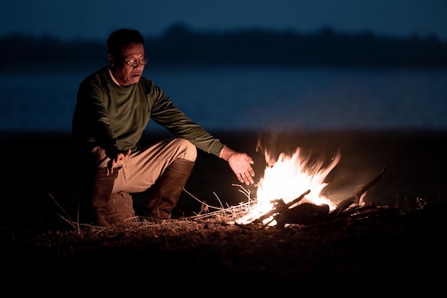 Free photo senior man in glasses sitting near lake warming his hands with the flames of a bonfire at cold weather in early moring, copy space