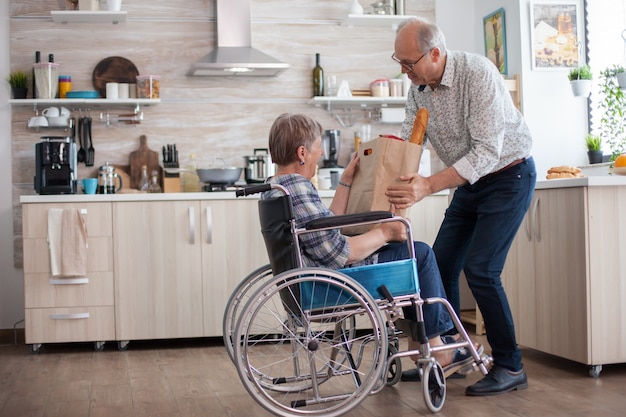 Free photo senior man helping his wife by taking grocery paper bag from her. mature people with fresh vegetables from market. living with disabled person with walking disabilities