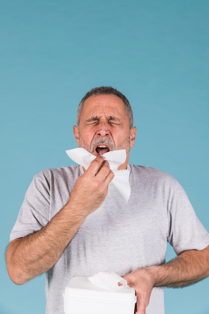 Senior man holding tissue paper about to sneeze on blue background