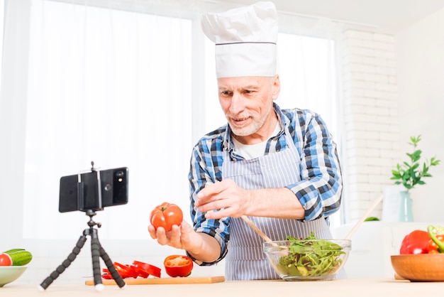 Free photo senior man making video call on mobile phone showing heirloom tomato while preparing salad