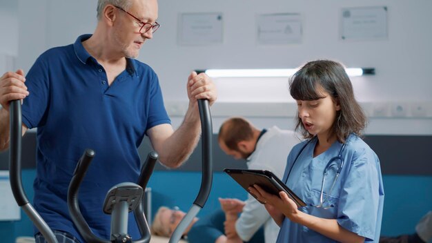 Senior man and medical nurse talking about osteopathy recovery, patient using stationary electrical bike to increase mobility. Old person attending rehabilitation procedure to heal.