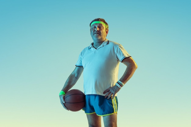 Senior man playing basketball on gradient background in neon light. Caucasian male model in great shape stays active, sportive. 