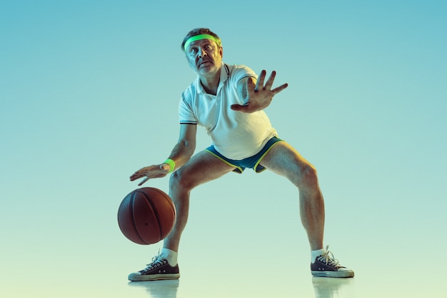 Senior man playing basketball on gradient wall in neon light. Caucasian male model in great shape stays active, sportive. Concept of sport, activity, movement, wellbeing, healthy lifestyle.