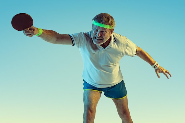 Free Photo senior man playing table tennis on gradient background in neon light. caucasian male model in great shape stays active, sportive. 